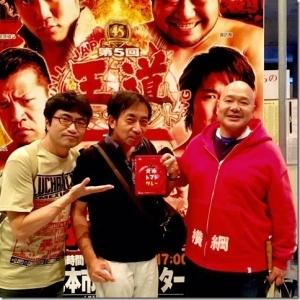 全日本プロレスのポスターの前で北本カレーを持って記念撮影をしている3名の男性の写真